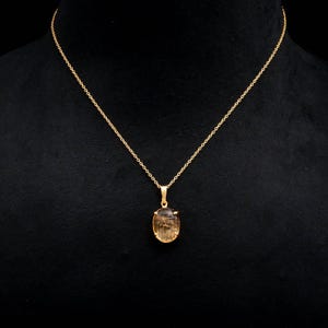 May include: A gold-colored necklace with a pendant featuring a light brown, oval-shaped gemstone. The chain is delicate, and the pendant is suspended from a small loop. The background is black, highlighting the jewelry.