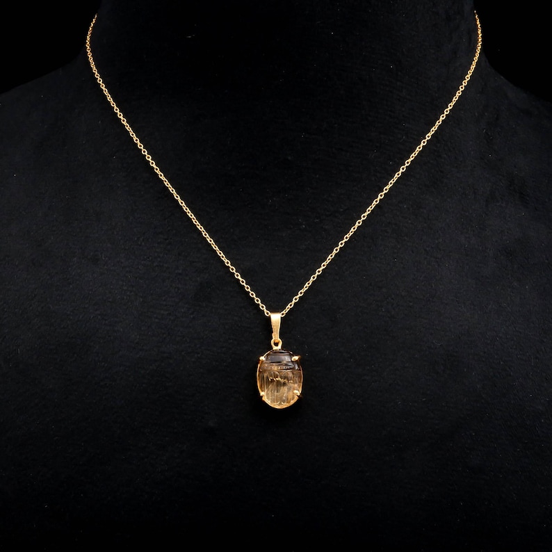 May include: A gold-colored necklace with a pendant featuring a light brown, oval-shaped gemstone. The chain is delicate, and the pendant is suspended from a small loop. The background is black, highlighting the jewelry.