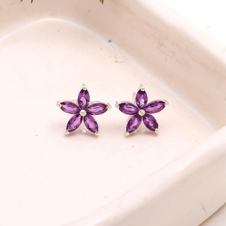 May include: A pair of flower-shaped stud earrings. Each earring features five purple gemstone petals set in a silver-colored metal. The earrings are displayed on a white surface.