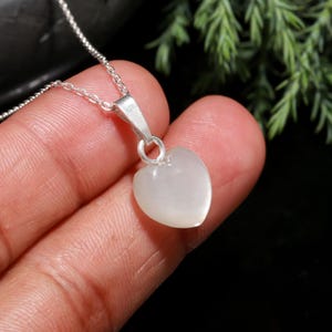 May include: A silver necklace with a heart-shaped pendant. The pendant is a pale, translucent color, and the chain is delicate. The necklace is displayed against a dark background, highlighting the pendant's shape and color.