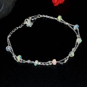 以下が含まれることがあります： 繊細なシルバーのブレスレット。2本の細いチェーンで構成され、小さな虹色のオパールビーズと小さなシルバービーズが飾られています。留め金には花の形のチャームが付いています。ブレスレットは暗い背景に表示されています。