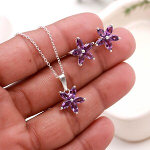May include: A silver necklace and matching earrings set. The pendant and earrings feature a flower design with six purple petals. The jewelry is set against a neutral background.
