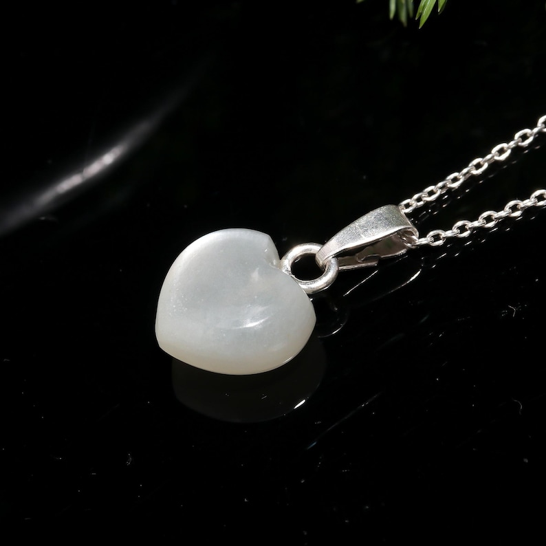 May include: A heart-shaped pendant necklace with a silver chain. The pendant is a pale, translucent color, and the chain is made of small, linked silver pieces. The necklace is displayed against a black background.