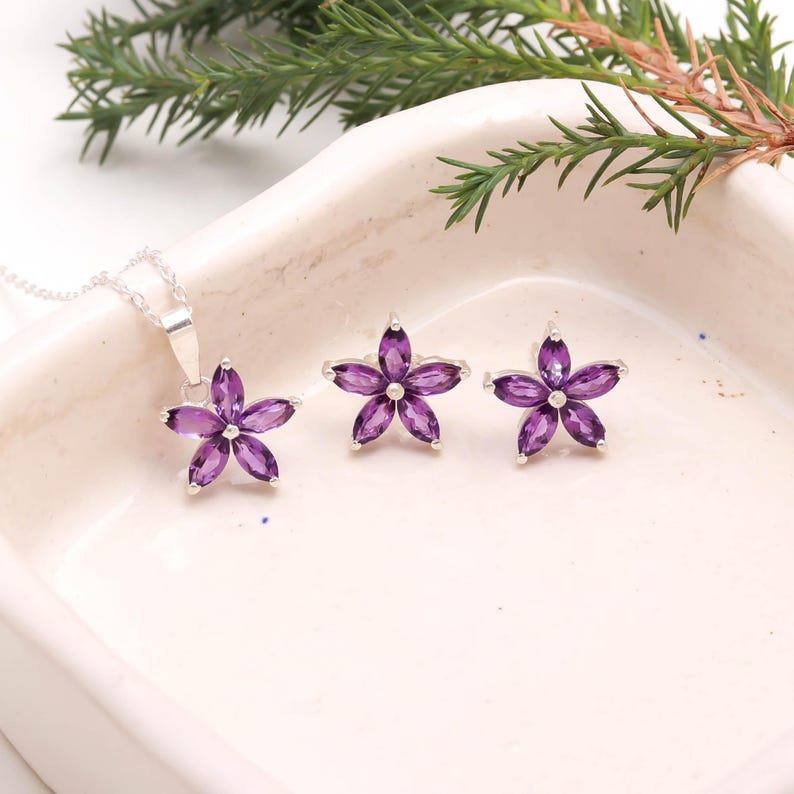 May include: A jewelry set featuring a pendant necklace and two flower-shaped earrings. The pendant and earrings are adorned with vibrant purple gemstones set in a silver metal. The floral design adds a touch of elegance.