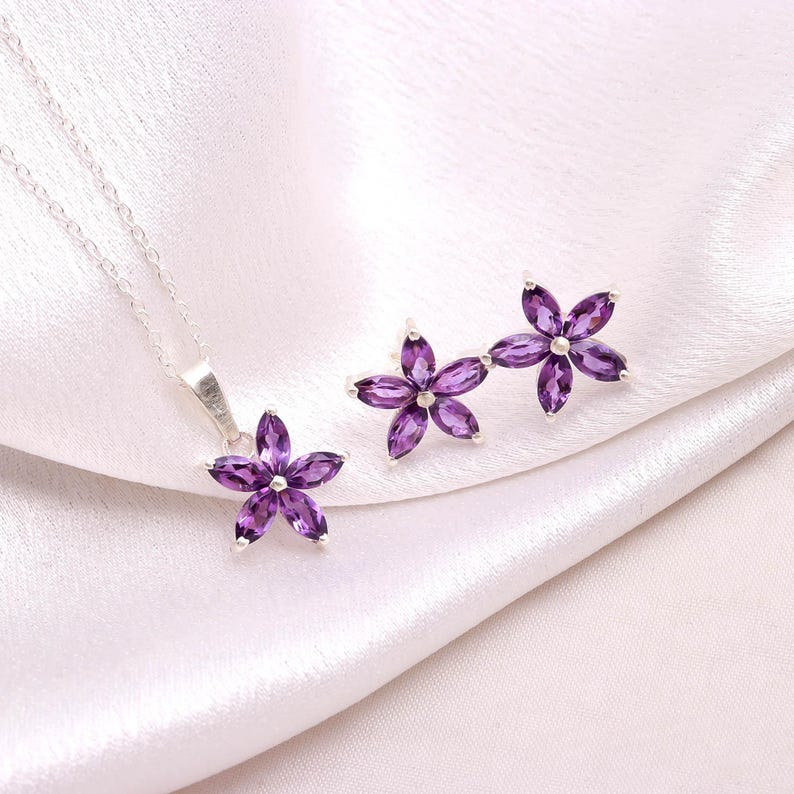 May include: A jewelry set featuring a silver necklace and earrings. The necklace has a pendant and the earrings are studs, all shaped like five-petal flowers. Each petal is a deep purple gemstone, set in silver, against a white satin background.