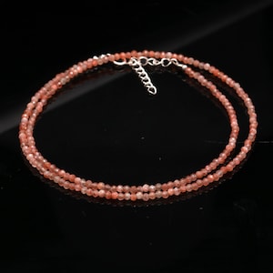 May include: A delicate necklace composed of small, faceted, round, peach-colored sunstone beads. The strand features a silver-toned metal lobster claw clasp and an adjustable chain link extender, allowing for variations in the length of the necklace.