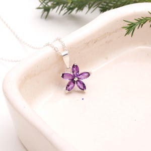 May include: A silver necklace with a pendant shaped like a flower. The flower pendant is made of six purple gemstones. The necklace is displayed on a white surface with green foliage in the background.
