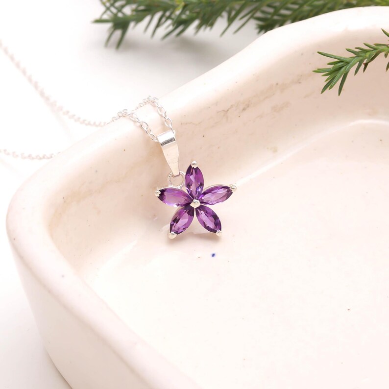 May include: A silver necklace with a pendant shaped like a flower. The flower pendant is made of six purple gemstones. The necklace is displayed on a white surface with green foliage in the background.
