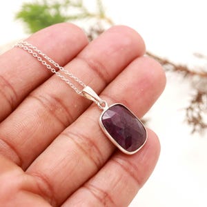 May include: A silver necklace featuring a rectangular, faceted deep red ruby pendant set in a smooth metal bezel. The pendant hangs from a delicate, thin silver chain, showcasing a polished finish and a dark, saturated gemstone color.