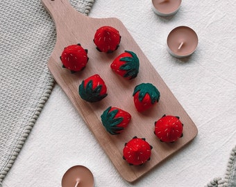 Può includere: Un tagliere di legno con otto fragole di feltro, ciascuna con una foglia verde, disposte in cerchio. Quattro piccole candele rotonde di colore marrone chiaro sono sparse intorno al tagliere.