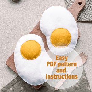 Puede incluir: Dos huevos de fieltro, uno blanco y otro amarillo, sobre una tabla de cortar de madera. Los huevos están rodeados de velas y un tenedor de madera. El texto "Easy PDF pattern and instructions" está escrito en un círculo blanco.