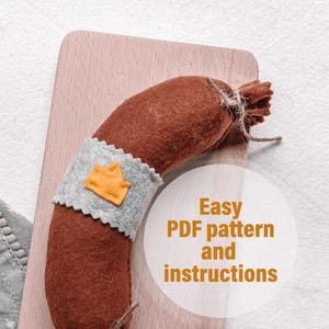 Könnte beinhalten: Eine braune Filz-Wurst mit einem grauen Filzband und einer gelben Filzkrone auf einem Holzbrett. Der Text "Easy PDF pattern and instructions" ist in Weiß auf einem weißen Kreis geschrieben.