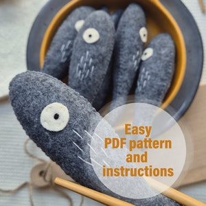 May include: A close-up of a gray felt fish toy with a white eye and black pupil. The fish is lying on a wooden surface with a pair of chopsticks and a bowl of gray felt fish toys in the background. The text "Easy PDF pattern and instructions" is written in a white circle on the image.