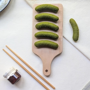 May include: A wooden cutting board with five green felt pickles on it. There are two jars of jam and a pair of chopsticks on a white surface.