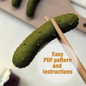May include: A close-up of a green felt pickle being held by chopsticks. The pickle is on a wooden surface with other felt pickles. The text "Easy PDF pattern and instructions" is on a white circle.