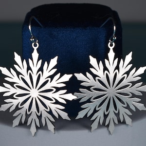 Könnte beinhalten: Ein Paar silberne Schneeflockenohrringe mit filigranen Ausschnitten. Die Ohrringe hängen an einem blauen Samt Hintergrund.
