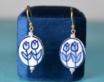 May include: Oval-shaped earrings with a white base and blue tulip design. The earrings have gold-colored hooks and small pearl accents. The earrings are displayed against a dark blue velvet background.
