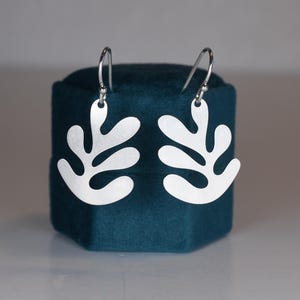 Puede incluir: Un par de pendientes plateados con forma de hoja. Los pendientes se exhiben en una caja de joyer&iacute;a de terciopelo azul verdoso. Los pendientes tienen un dise&ntilde;o sencillo y moderno con un acabado mate y cierre de gancho.