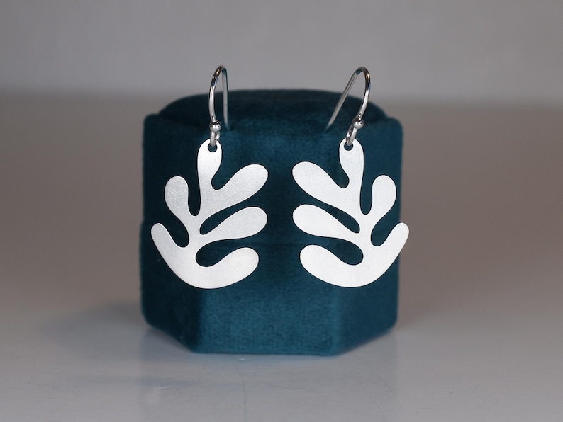 Puede incluir: Un par de pendientes plateados con forma de hoja. Los pendientes se exhiben en una caja de joyer&iacute;a de terciopelo azul verdoso. Los pendientes tienen un dise&ntilde;o sencillo y moderno con un acabado mate y cierre de gancho.