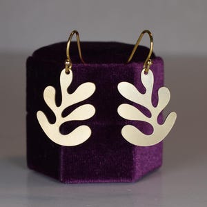 Puede incluir: Un par de pendientes dorados con un dise&ntilde;o en forma de hoja. Los pendientes se exhiben en una caja de terciopelo morado. Los pendientes tienen un cierre de gancho.
