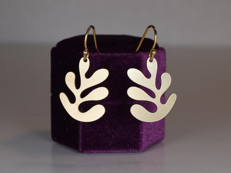 Puede incluir: Un par de pendientes dorados con un dise&ntilde;o en forma de hoja. Los pendientes se exhiben en una caja de terciopelo morado. Los pendientes tienen un cierre de gancho.
