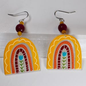 May include: A pair of yellow rainbow earrings with a red, orange, and yellow rainbow design. The earrings have a burgundy bead and a silver hook.