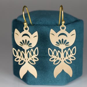 May include: A pair of gold-colored, floral-shaped earrings. Each earring features a stylized flower design with a crescent moon shape at the top, a detailed center, and a fish-tail bottom. The earrings are displayed on a teal-colored surface.