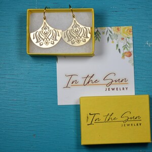 Puede incluir: Pendientes dorados en forma de abanico con intrincados dise&ntilde;os calados, colocados en una caja amarilla. La caja est&aacute; sobre una superficie azul junto a una tarjeta con detalles florales y el texto "In the Sun JEWELRY". Tambi&eacute;n hay otra caja amarilla con el mismo texto.