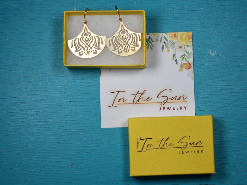 Puede incluir: Pendientes dorados en forma de abanico con intrincados dise&ntilde;os calados, colocados en una caja amarilla. La caja est&aacute; sobre una superficie azul junto a una tarjeta con detalles florales y el texto "In the Sun JEWELRY". Tambi&eacute;n hay otra caja amarilla con el mismo texto.