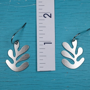 Puede incluir: Un par de pendientes plateados con forma de hoja. Los pendientes miden aproximadamente 2,5 cm de largo. Cuelgan de un gancho simple. El fondo es una superficie de madera turquesa desgastada.