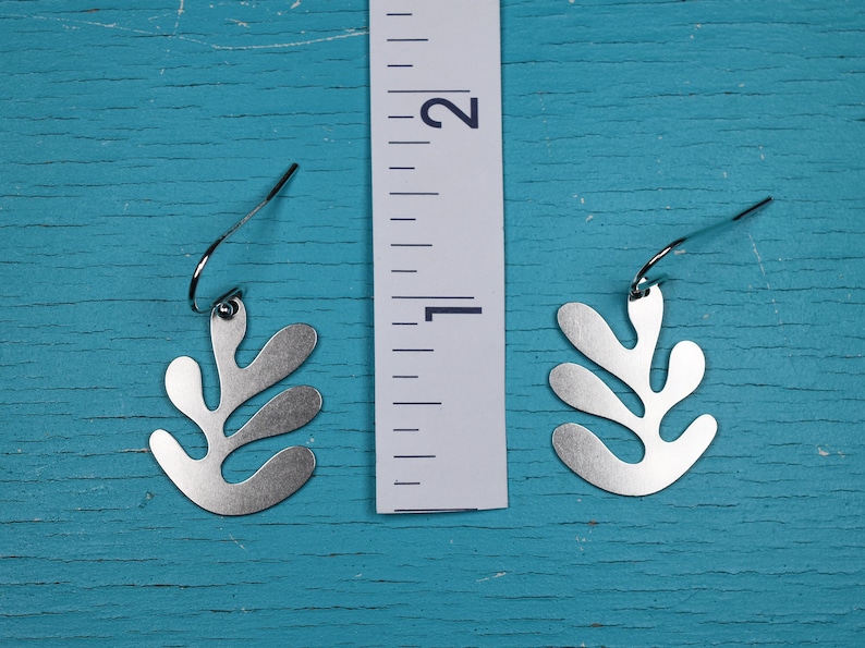 Puede incluir: Un par de pendientes plateados con forma de hoja. Los pendientes miden aproximadamente 2,5 cm de largo. Cuelgan de un gancho simple. El fondo es una superficie de madera turquesa desgastada.