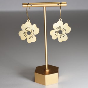 May include: Gold-colored flower-shaped earrings with a detailed center and delicate petals. The earrings are displayed on a gold-toned stand with a hexagonal base, set against a neutral background. The earrings are lightweight and suitable for everyday wear.