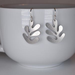 Puede incluir: Un par de pendientes plateados con dise&ntilde;o de hoja. Los pendientes cuelgan de ganchos plateados y se muestran dentro de una taza blanca. Los pendientes tienen un acabado mate y una forma org&aacute;nica &uacute;nica.