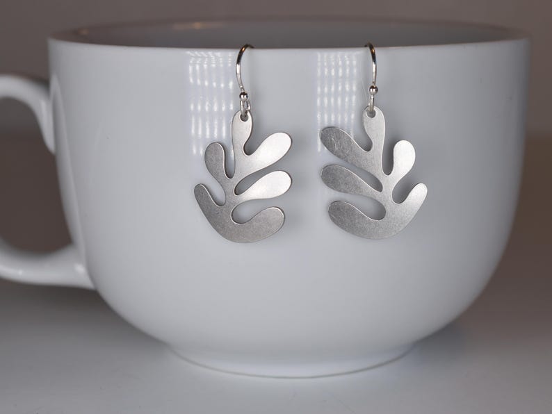 Puede incluir: Un par de pendientes plateados con dise&ntilde;o de hoja. Los pendientes cuelgan de ganchos plateados y se muestran dentro de una taza blanca. Los pendientes tienen un acabado mate y una forma org&aacute;nica &uacute;nica.