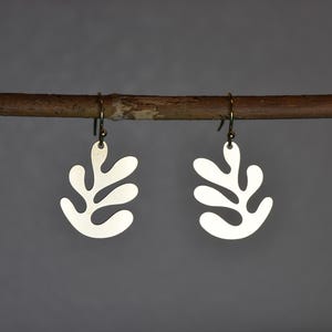 Puede incluir: Un par de pendientes plateados con forma de hoja colgando de una rama marr&oacute;n. Los pendientes tienen un dise&ntilde;o sencillo y moderno con un acabado mate. Los ganchos son de un color lat&oacute;n antiguo m&aacute;s oscuro. El fondo es gris neutro liso.