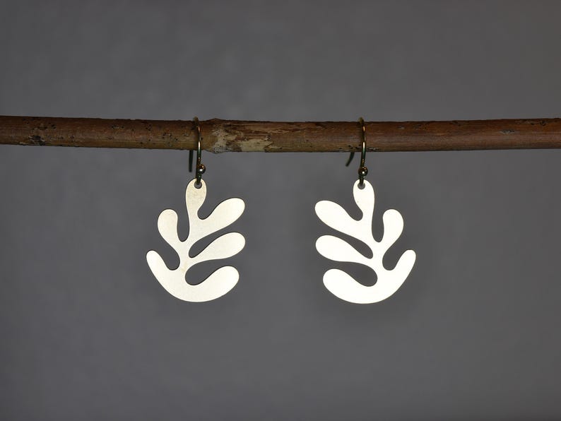Puede incluir: Un par de pendientes plateados con forma de hoja colgando de una rama marr&oacute;n. Los pendientes tienen un dise&ntilde;o sencillo y moderno con un acabado mate. Los ganchos son de un color lat&oacute;n antiguo m&aacute;s oscuro. El fondo es gris neutro liso.