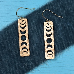 May include: A pair of gold-toned earrings with a hammered texture. Each earring features a rectangular shape with a cutout design of eight crescent moons.