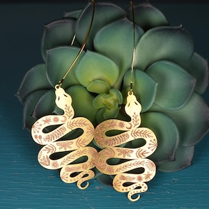 May include: A pair of gold-tone snake earrings with intricate floral and celestial designs. The earrings are hanging from a green succulent plant.