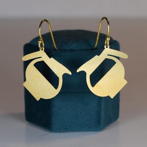 May include: A pair of gold-colored earrings with a unique, abstract design. The earrings are displayed on a dark teal velvet jewelry box. The earrings have a hook closure.