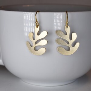 Puede incluir: Un par de pendientes dorados con un dise&ntilde;o en forma de hoja. Los pendientes cuelgan de ganchos dorados. Los pendientes se muestran dentro de una taza blanca.