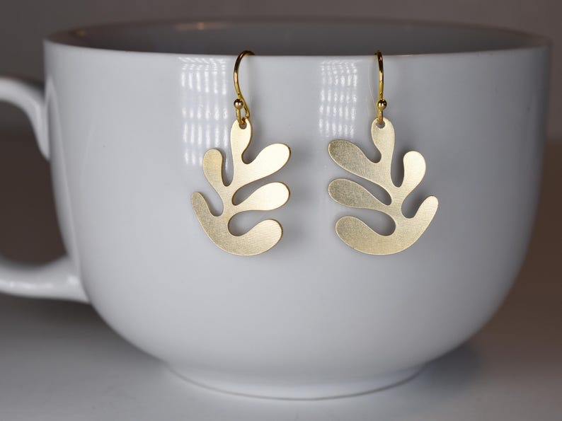 Puede incluir: Un par de pendientes dorados con un dise&ntilde;o en forma de hoja. Los pendientes cuelgan de ganchos dorados. Los pendientes se muestran dentro de una taza blanca.