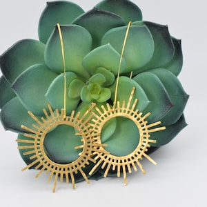 May include: A pair of gold-toned earrings with a sunburst design. The earrings are made of metal and have a circular shape with a delicate, openwork pattern. The earrings are hanging from a green succulent plant.