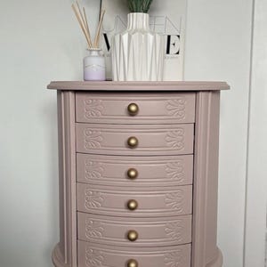 Op de afbeelding: Een mauvekleurige sieradenkast met zes lades, elk versierd met goudkleurige knoppen. De lades hebben een sierlijk, gesneden ontwerp. De kast heeft een gebogen basis en poten en wordt bekroond door een vaas met groen.