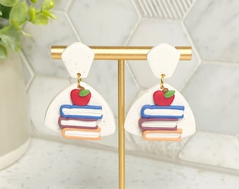 May include: A pair of white dangle earrings with a stack of books and a red apple on top. The earrings are on a gold stand.