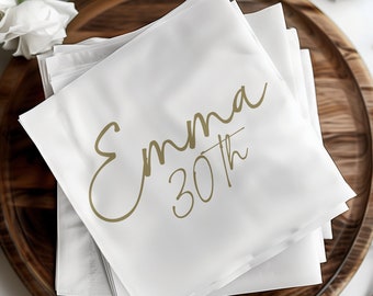 May include: A stack of white napkins with gold lettering that reads "Emma 30th".