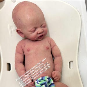 May include: A realistic newborn baby doll with pale skin, closed eyes, and subtle mottling, lying on a white plastic surface. A small blue, green, and white striped cloth covers the lower body. Text on the doll reads: Website: carolineheussdolls.com, carolinehdolls.etsy.com, CarolineHeussDolls1.Etsy.com, Instagram: @carolineheussdolls, Facebook: CarolineHeussDolls.