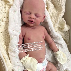 May include: A realistic newborn baby doll with pale skin, closed eyes, and a slightly open mouth. The doll wears a floral print diaper, pale yellow mittens, and light pink socks. Text reads: Website: carolineheussdolls.com carolinehdolls.etsy.com CarolineHeussDolls1.Etsy.com Instagram: @carolineheussdolls Facebook: CarolineHeussDolls