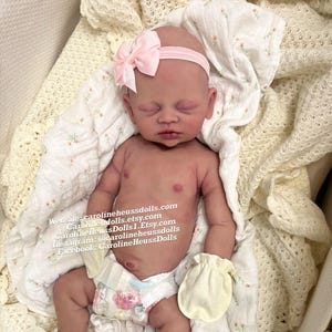 May include: A realistic newborn baby doll with closed eyes, wearing a light pink headband with a bow, pale yellow mittens, pink socks, and a diaper with a floral pattern. The text on the image reads: Website: carolineheussdolls.com, CarolineHeussDolls.Etsy.com, Instagram: @carolineheussdolls, Facebook: CarolineHeussDolls.