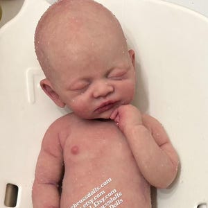 May include: A realistic silicone baby doll with closed eyes, wearing a blue and green striped towel, lying on its back in a white tub.