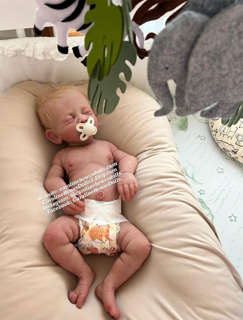 Op de afbeelding: Een realistische babypop met blond haar, die een luier en een speen draagt, ligt op een beige deken. De pop is omringd door een jungle-mobiel met een zebra, bladeren en een olifant.